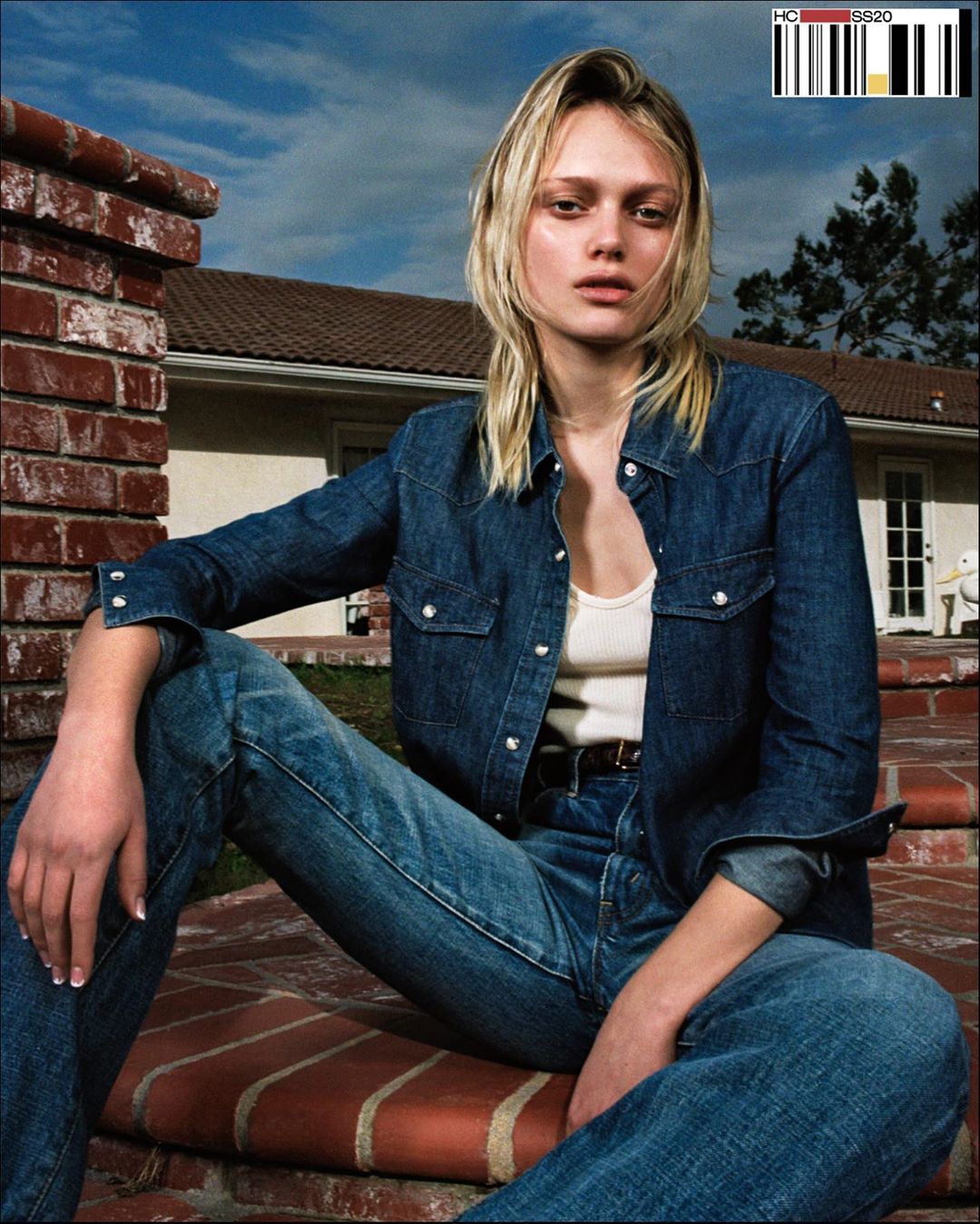 A young adult woman with blonde hair poses on brick steps wearing a denim shirt and jeans with a house and blue sky background.