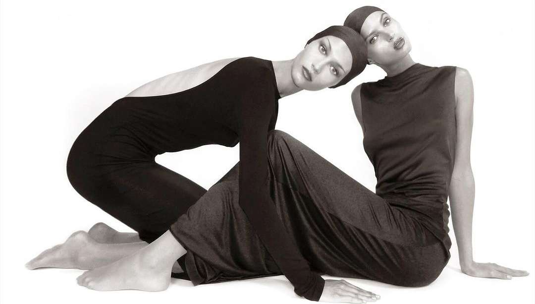 Two fashion models pose in matching black head coverings and fitted long dresses in a studio lit monochrome fashion editorial shot.