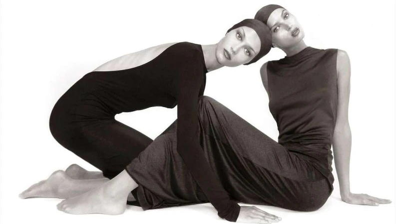 Two fashion models pose in matching black head coverings and fitted long dresses in a studio lit monochrome fashion editorial shot.
