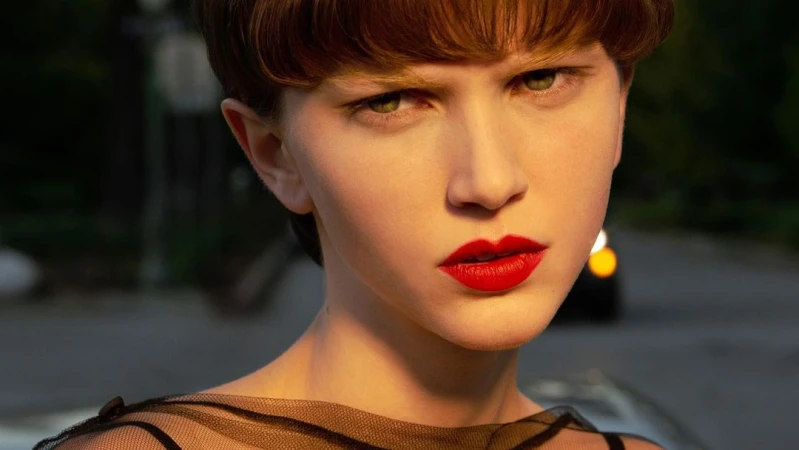 Close-up portrait of a model with short brown hair, red lipstick, and sheer black top outdoors at sunset by Hugo Comte.