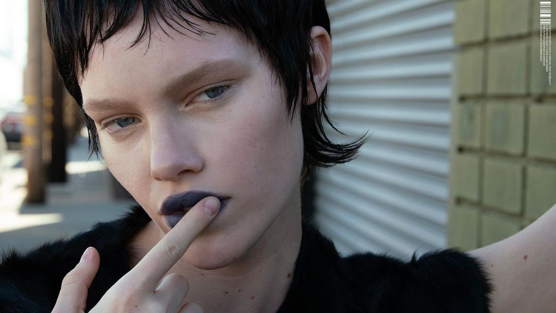 Close-up of a model with dark, textured lipstick and dark hair, posed against a background of vertical siding and weathered brick.