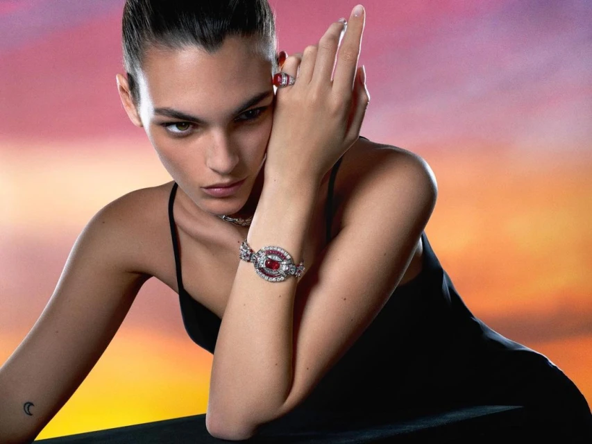 Adult female model poses indoors, wearing a black top and jewelry, against a blended pink-and-orange gradient background.