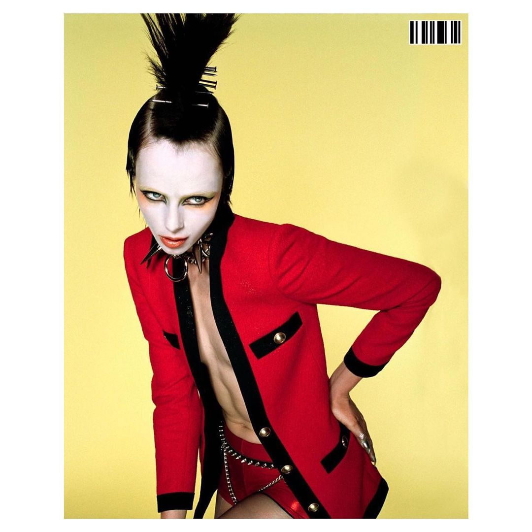 Edgy fashion shot of a slender woman with extreme pale makeup, spiky hair, wearing a red blazer and studded collar against a yellow backdrop.