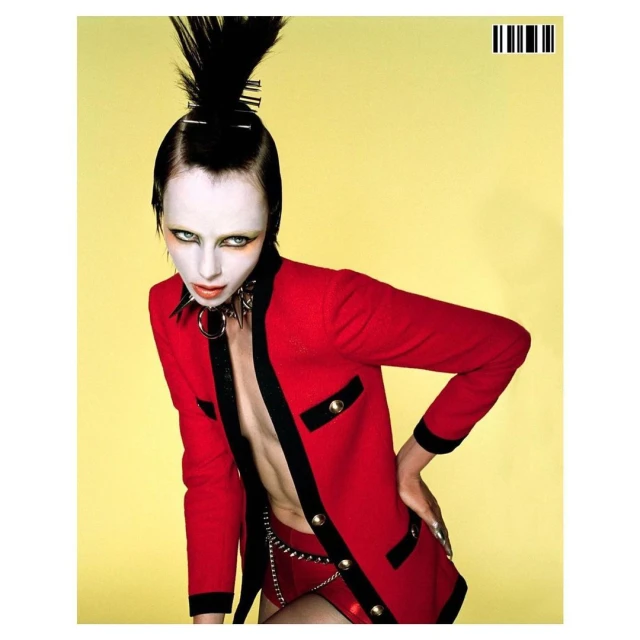 Edgy fashion shot of a slender woman with extreme pale makeup, spiky hair, wearing a red blazer and studded collar against a yellow backdrop.