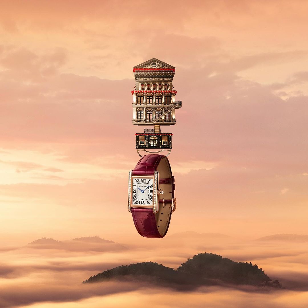 A stylish wristwatch is displayed with a building model on top, suspended against a backdrop of misty mountains and a soft orange sky.