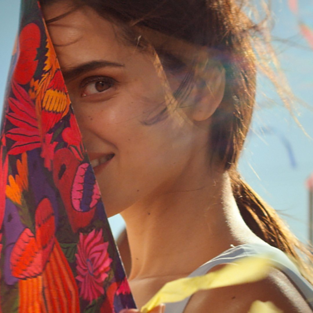 Portrait of a smiling woman with brown hair in a ponytail, holding a colourful floral-patterned fabric, set against a bright, clear sky.