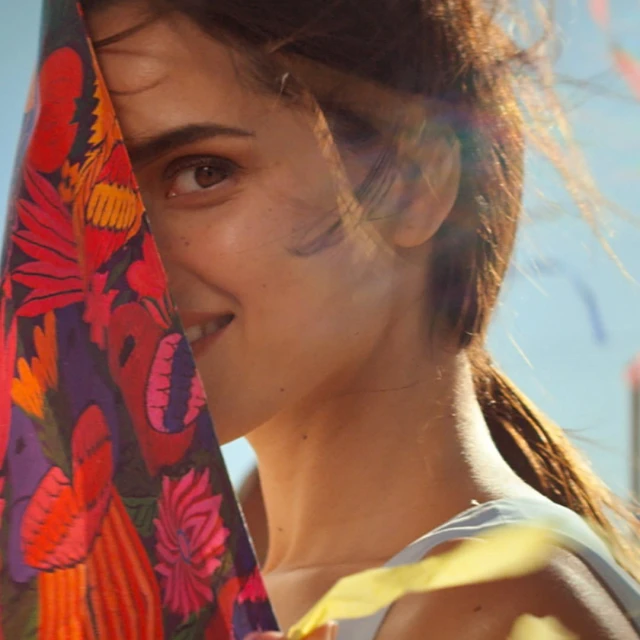 Portrait of a smiling woman with brown hair in a ponytail, holding a colourful floral-patterned fabric, set against a bright, clear sky.