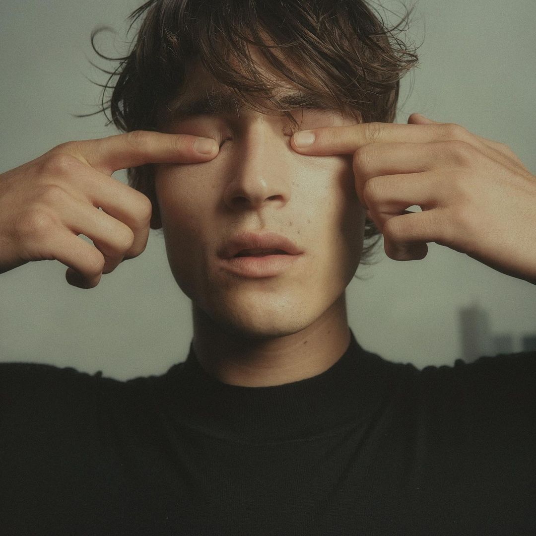 Portrait of a young adult male with fingers touching eyelids, wearing a black turtleneck shirt, with blurred buildings in the background.