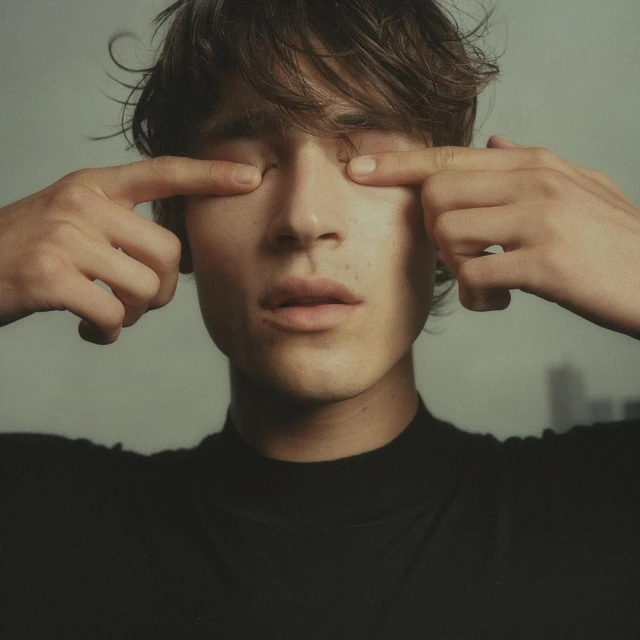 Portrait of a young adult male with fingers touching eyelids, wearing a black turtleneck shirt, with blurred buildings in the background.
