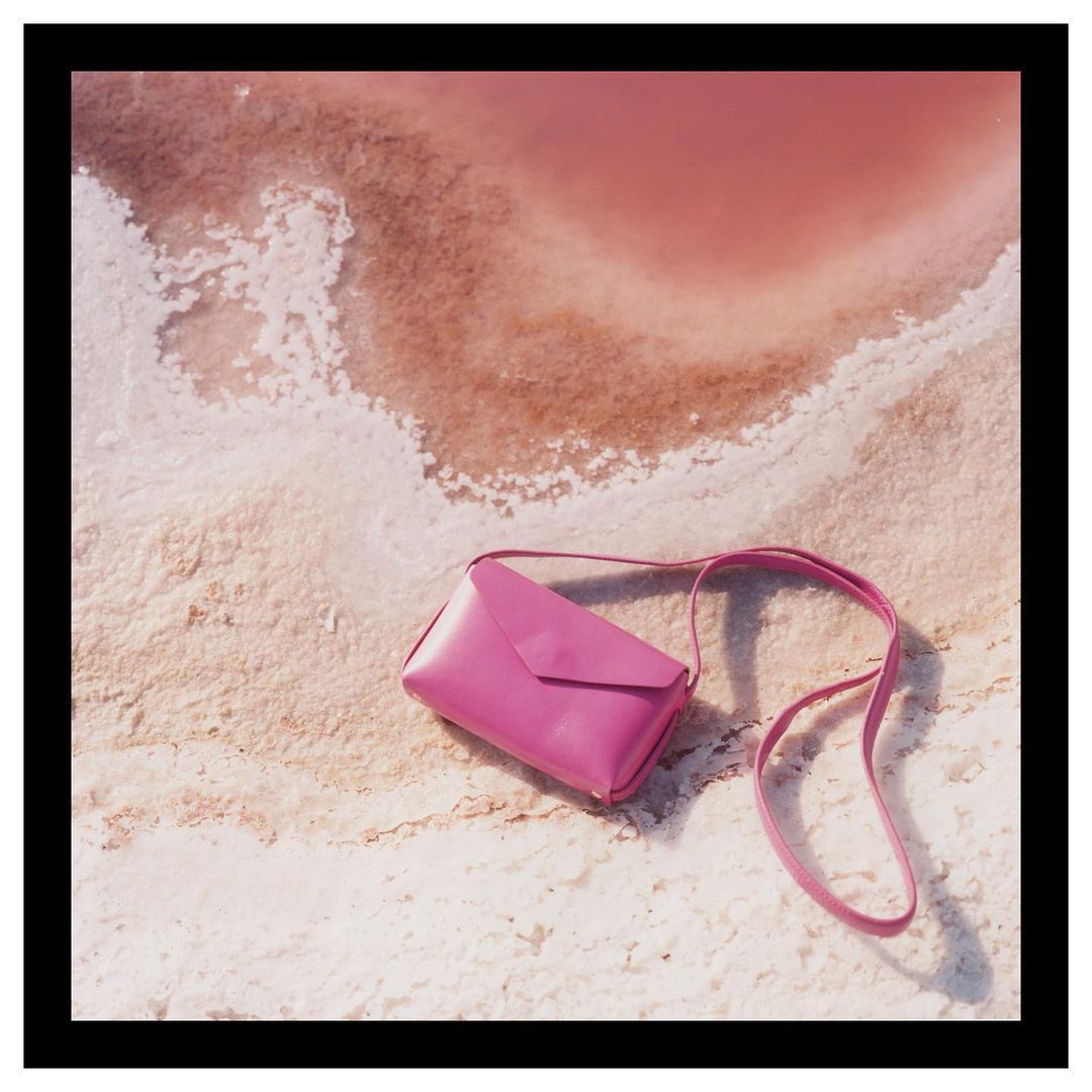 Pink leather envelope purse with shoulder strap, placed on textured sand near a pink lake or water body, fashion accessory still life.