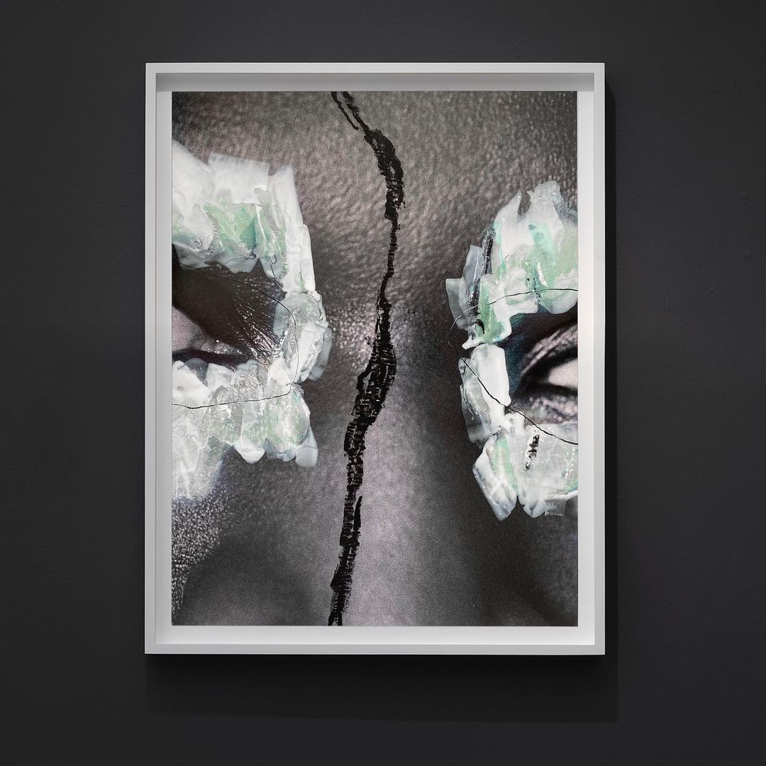 Conceptual close-up of a person's face with abstract, geometric makeup around the eyes and a central vertical line, framed and hanging on a wall.