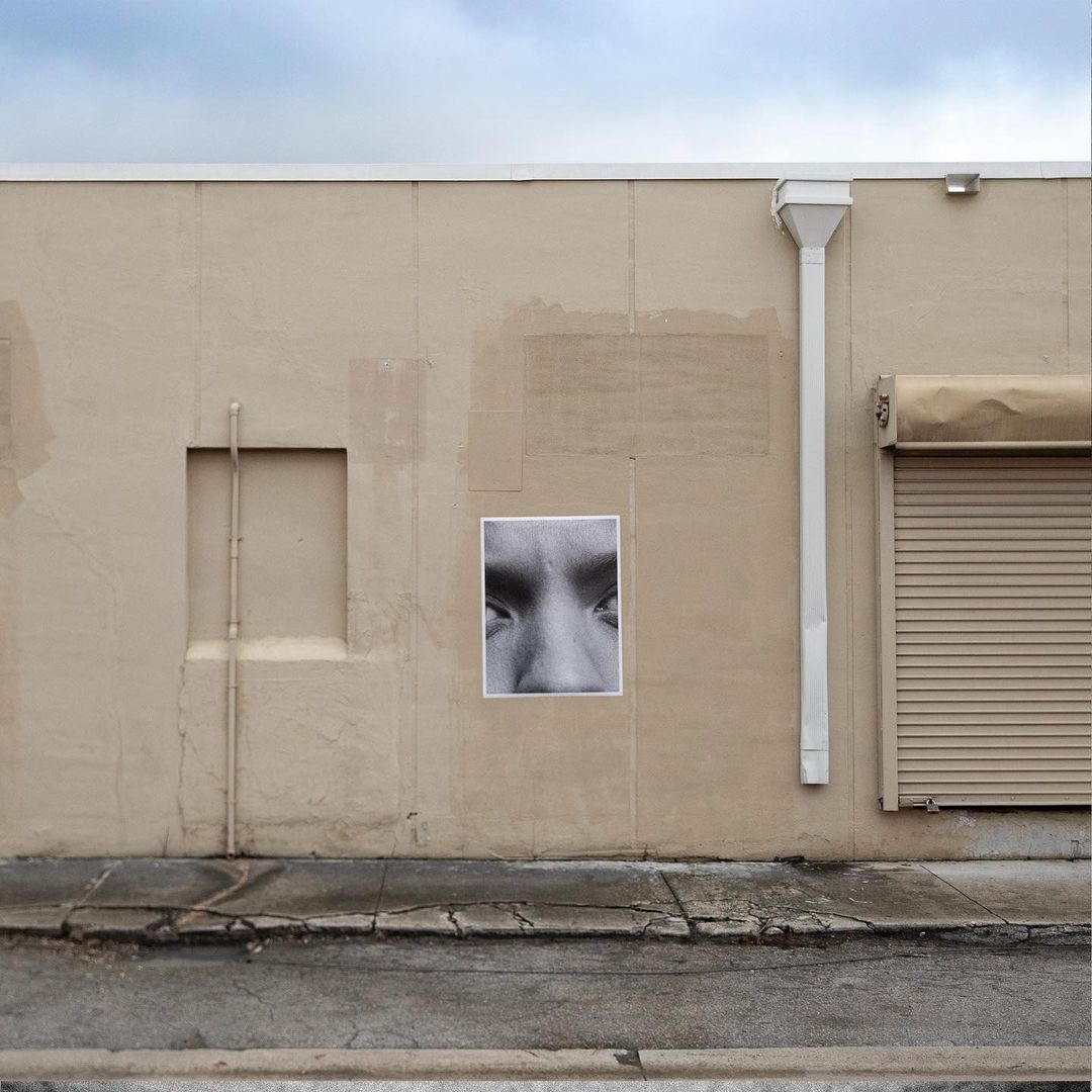 Monochrome image of human eyes and nose on a poster attached to a tan building with a partially-closed roll-up door and blue sky.