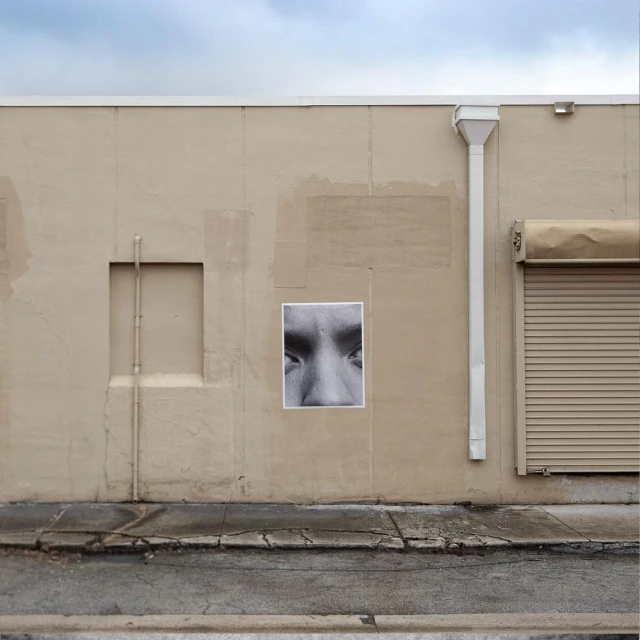 Monochrome image of human eyes and nose on a poster attached to a tan building with a partially-closed roll-up door and blue sky.