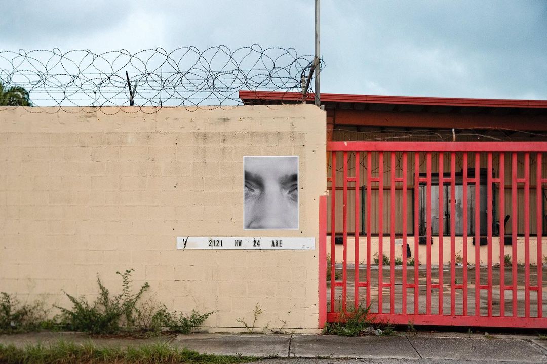 Street art on a weathered wall features a large eye poster, barbed wire, and red metal gate. Cloudy skies above an urban landscape.