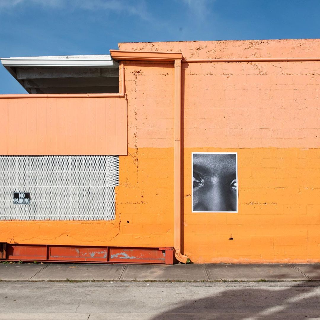 Eye-level shot of a black and white photographic print featuring human eyes on a peach and orange building with a 'No Parking' sign.