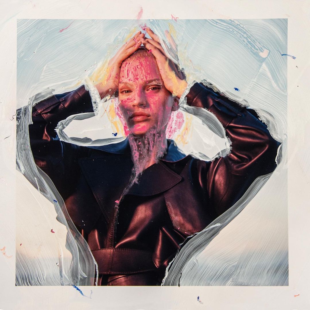 Artistic portrait of a person with pink coloration on their head and face, wearing a black jacket, set against a textured white background.