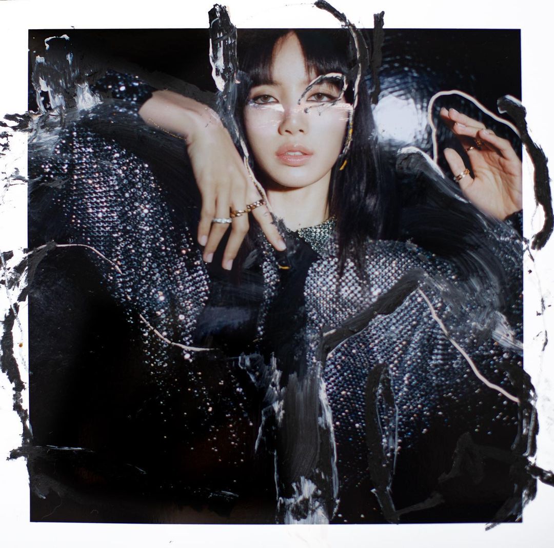 A woman with dark hair in a shiny jacket with paint streaks, fashion photography, studio shot, dark colors, rings and necklace.