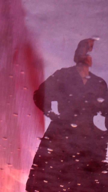 A woman is reflected in a wet surface with pink and purple hues, wearing a dark coat, creating a blurry and artistic composition.