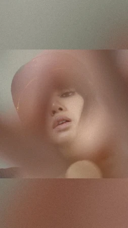 Portrait of a person with light skin wearing a hat, partially obscured by blurry foreground, shot with a low angle and diffused lighting.