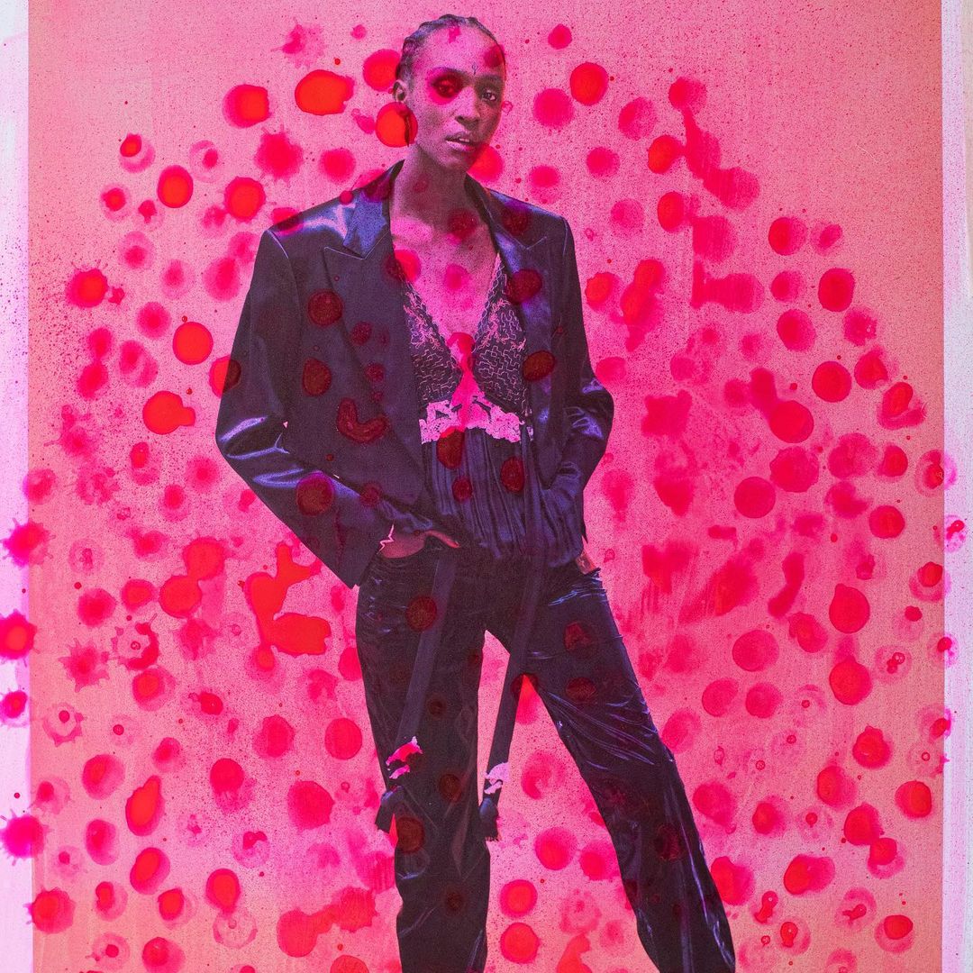 Fashion image of an adult woman in a dark blazer and pantsuit, posing in front of a pink backdrop with red dots. Indoor setting.