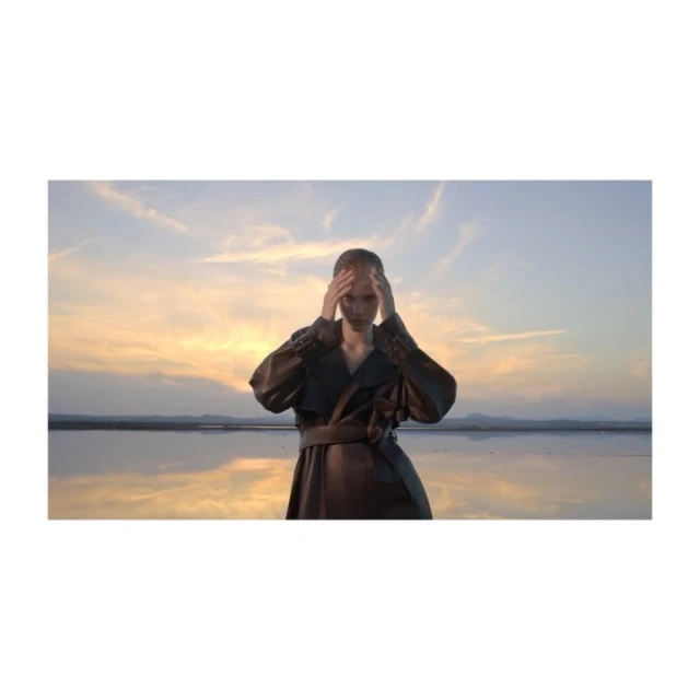 Woman in a long black coat with her hands on her head stands in a shallow body of water with a golden sunset in the background.