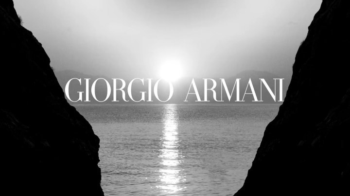 Black and white photo of a sea view framed by dark cliffs, featuring the text 'GIORGIO ARMANI' over the water.