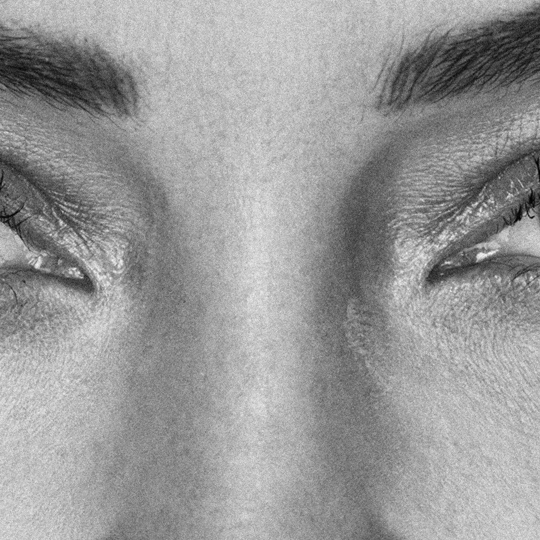 Close-up, black and white portrait captures the intense gaze and facial details of a woman, highlighting her eyebrows, eyes, and skin texture.
