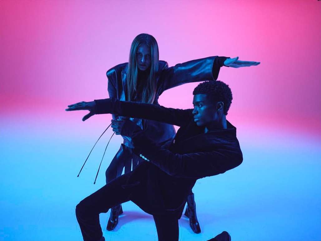 Two fashion models strike a pose in a photo studio with blue and pink lighting. One African-American male and one light-skinned female are featured.