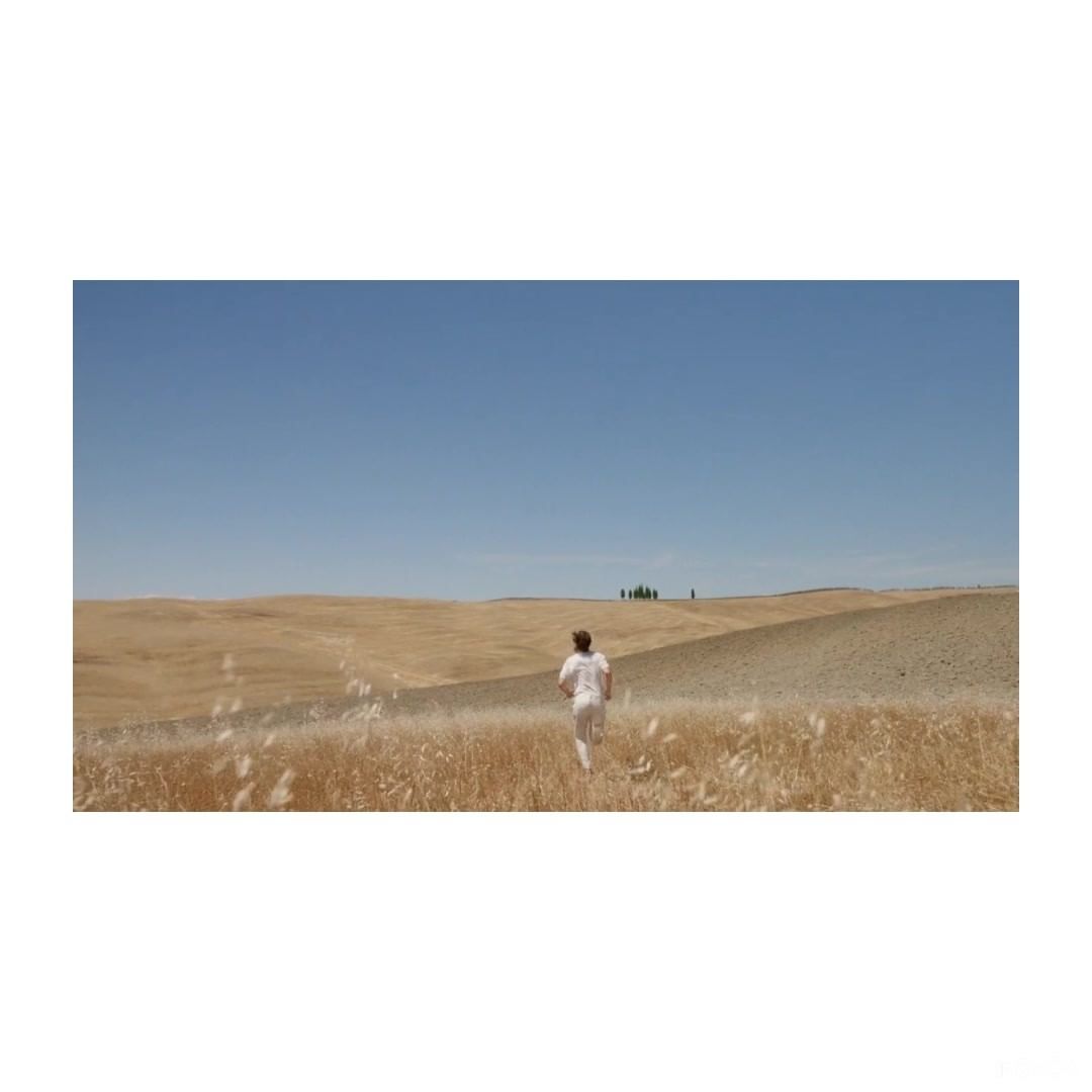 A woman runs through a dry field under a clear blue sky in a sun-drenched rural landscape, wearing casual clothing and seemingly enjoying the solitude.