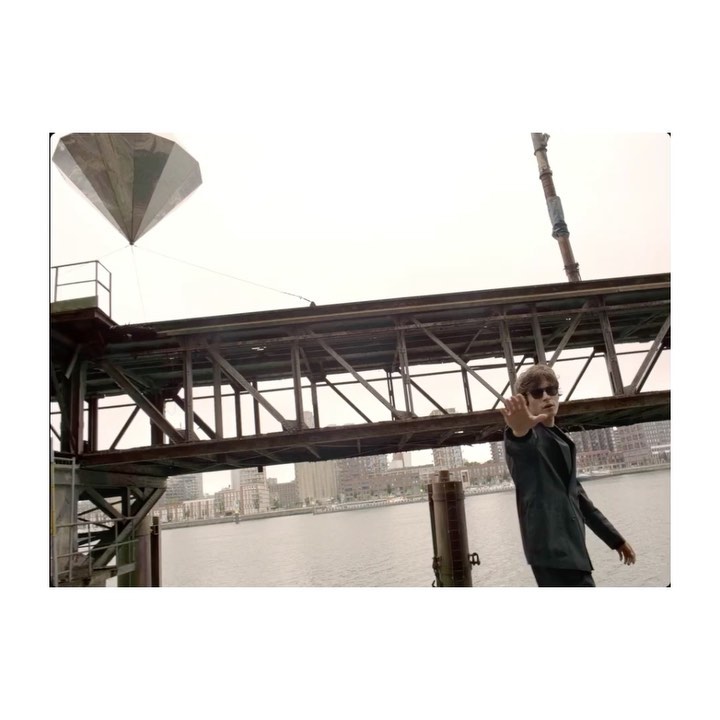 Man in suit poses on pier with large diamond sculpture, river, city skyline, and cloudy sky in background, medium outdoor shot.
