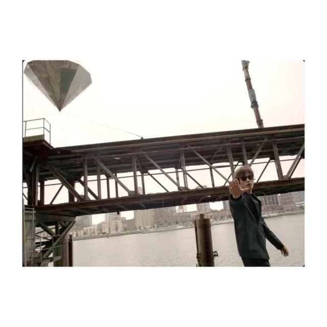 Man in suit poses on pier with large diamond sculpture, river, city skyline, and cloudy sky in background, medium outdoor shot.