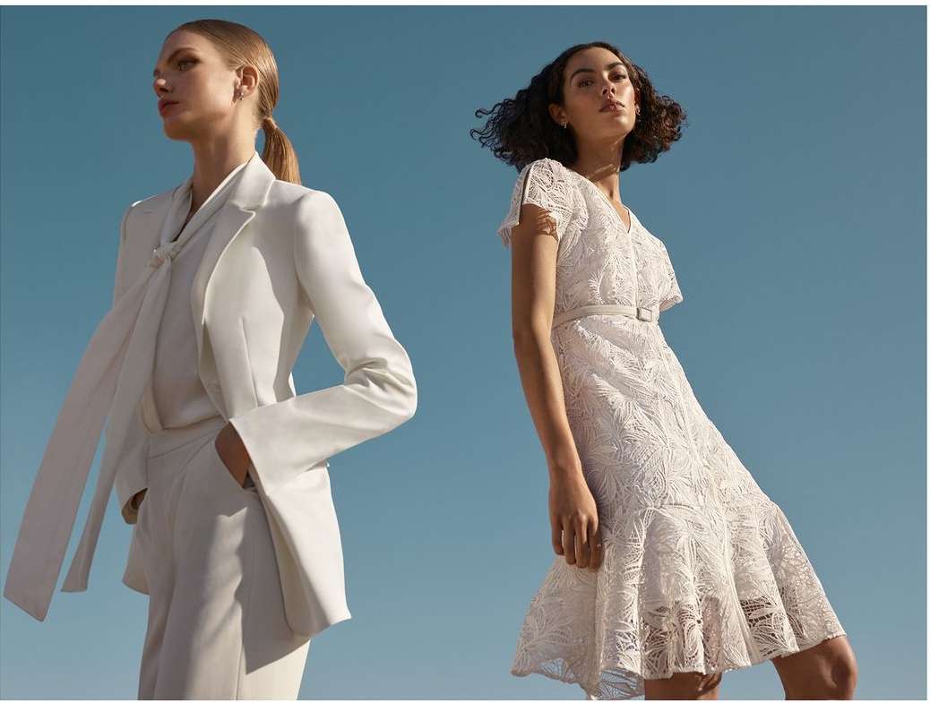 Two women pose against a clear blue sky, one in a white blazer and shorts set, the other in a white lace mini-dress, in a fashion setting.