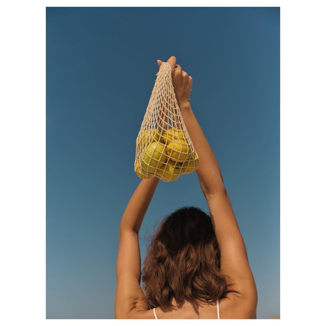 Woman holding a mesh bag filled with green apples against a clear blue sky, shot from a low angle, emphasizing the fruit and open sky.