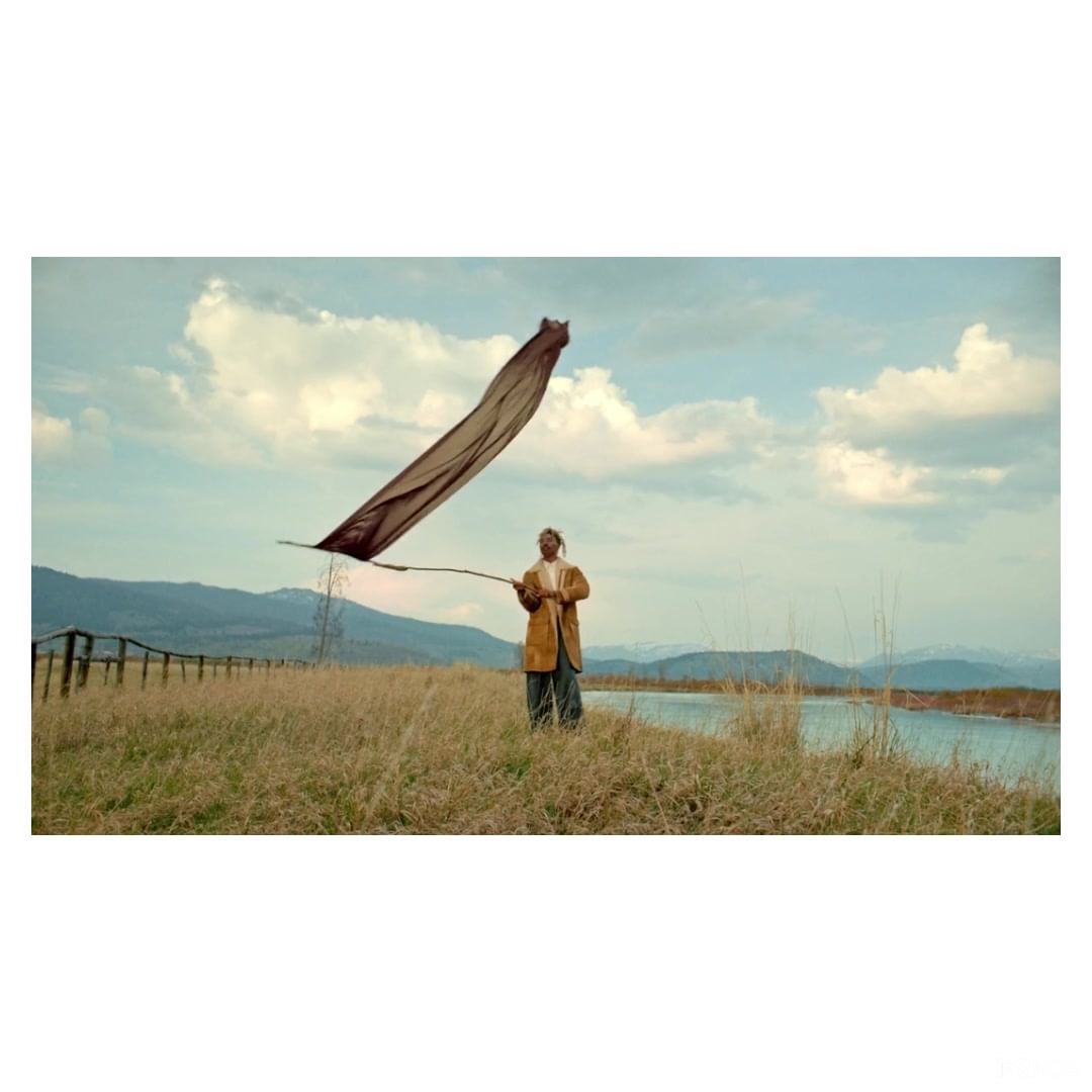 Adult stands on a field near a lake holding a stick with a fabric blowing in the wind against a blue sky with clouds and mountains.
