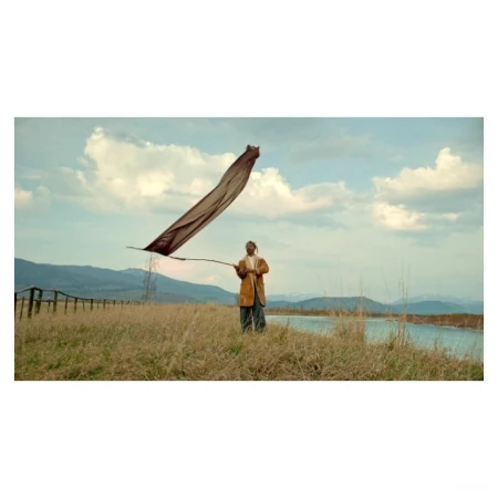 Adult stands on a field near a lake holding a stick with a fabric blowing in the wind against a blue sky with clouds and mountains.