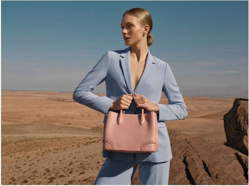 Woman in a light blue suit stands on a rocky terrain, holding a pink purse against a desert and sky background in a fashion shot.