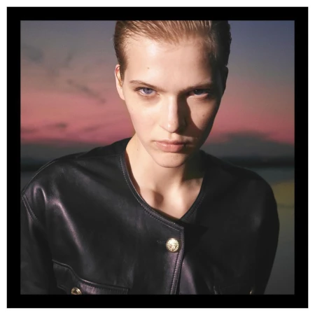 Close-up portrait of a model with slicked-back hair wearing a dark leather jacket against a sunset sky.