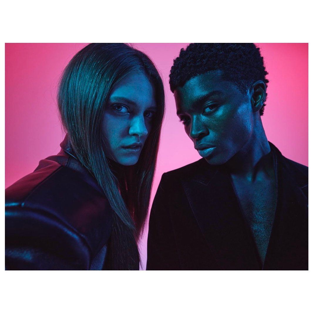 Fashion shot of a young adult couple in a photo studio with pink and blue lighting wearing black clothing. Medium shot, looking at camera.