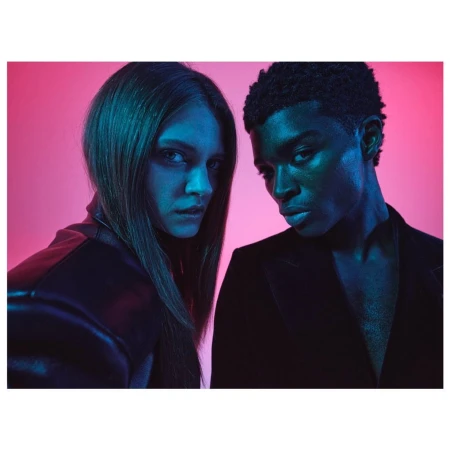 Fashion shot of a young adult couple in a photo studio with pink and blue lighting wearing black clothing. Medium shot, looking at camera.