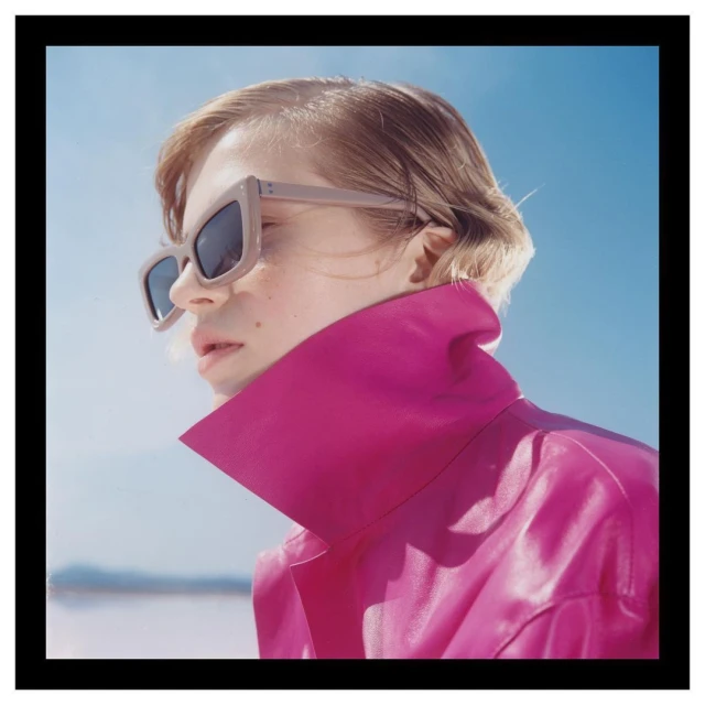 Close-up of a fair-skinned young adult wearing sunglasses and a magenta jacket with a high collar against a blue sky background.