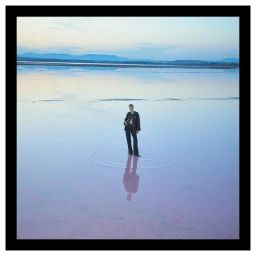 A model standing in shallow water reflecting the sky, wearing a dark jacket and trousers by Hunter and Gatti.