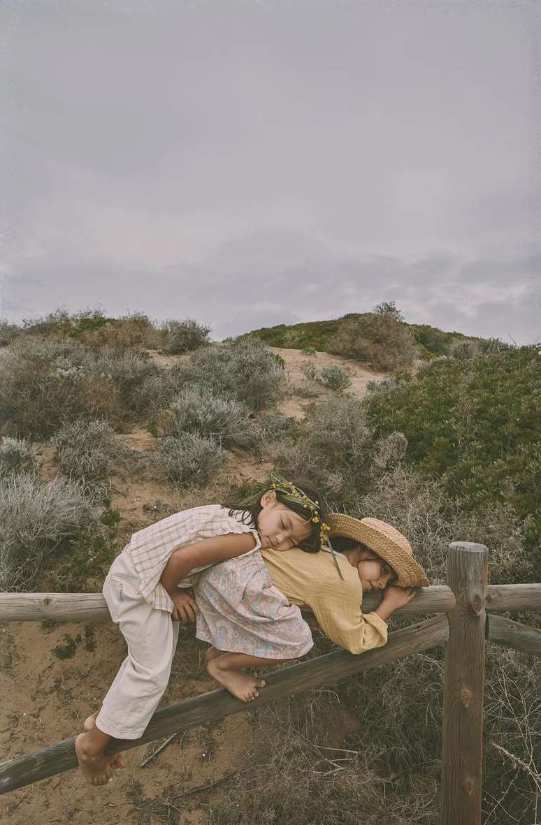 Two young children in vintage clothing are nestled together on a wooden fence against a natural landscape with a cloudy sky backdrop.