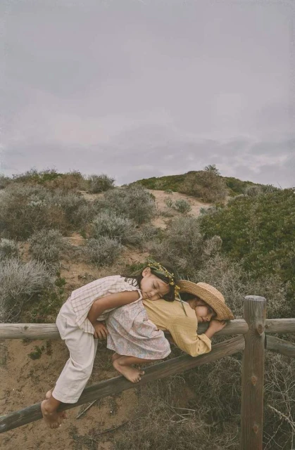 Two young children in vintage clothing are nestled together on a wooden fence against a natural landscape with a cloudy sky backdrop.