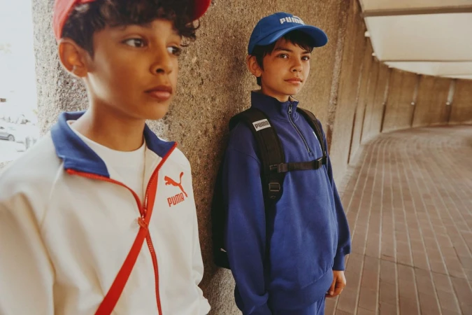 Two young boys wearing Puma branded activewear, one in a white jacket with red trim and the other in blue, stand against a textured wall.
