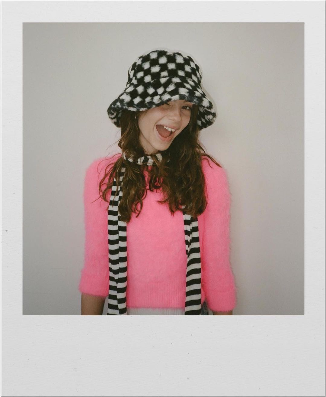Portrait of smiling young woman wearing a black and white checkered fuzzy bucket hat and bright pink sweater.