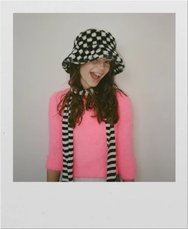 Portrait of smiling young woman wearing a black and white checkered fuzzy bucket hat and bright pink sweater.