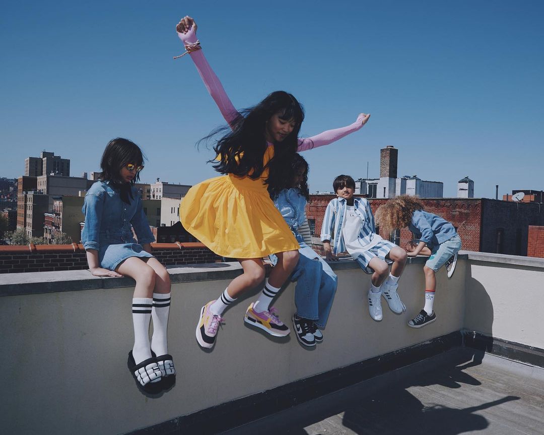 Group of children playing on a rooftop with one girl mid jump wearing a bright yellow dress under a clear blue sky