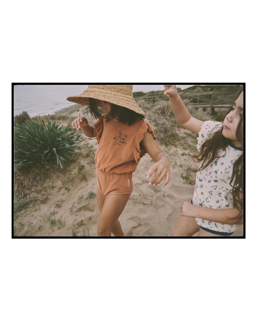 Two children pose on a sandy beach in a vintage-toned shot. One child wears a straw hat and a matching brown romper. Ocean visible.