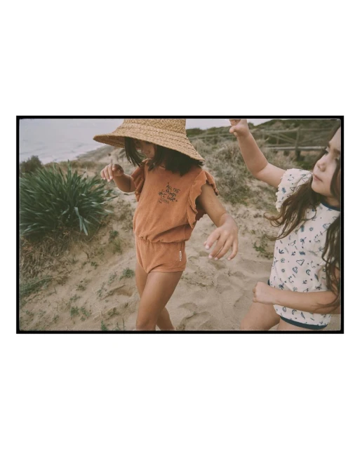 Two children pose on a sandy beach in a vintage-toned shot. One child wears a straw hat and a matching brown romper. Ocean visible.