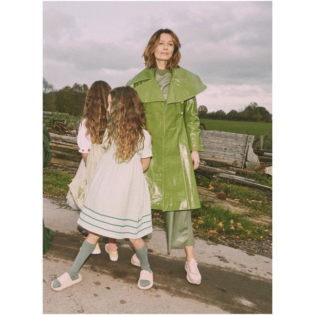A mother and two daughters wearing green coats and dresses pose on a rural road with a cloudy sky and farm in the background.